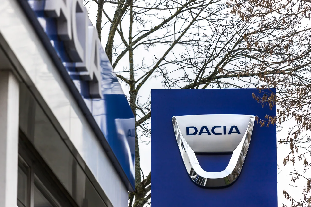 Sigla Dacia pe un panou albastru, în fața unei clădiri, lângă un copac desfrunzit