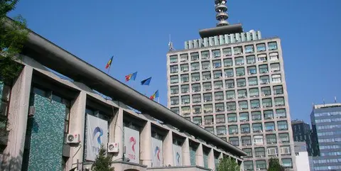 Clădire-turn de birouri cu antenă pe acoperiș, lângă o clădire mai joasă cu drapele, sub cer senin