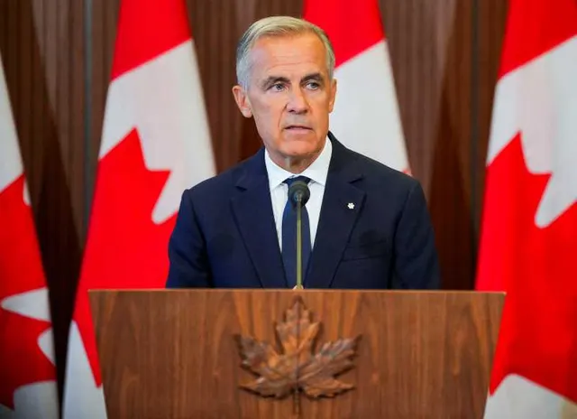 Bărbat cu părul grizonat, la un pupitru de lemn cu frunza de arțar, în fața drapelelor Canadei