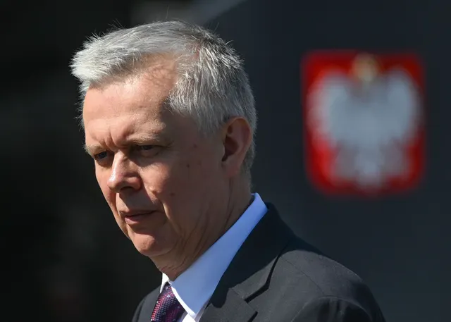 Bărbat cu părul grizonat, în costum, lângă stema Poloniei cu vulturul alb