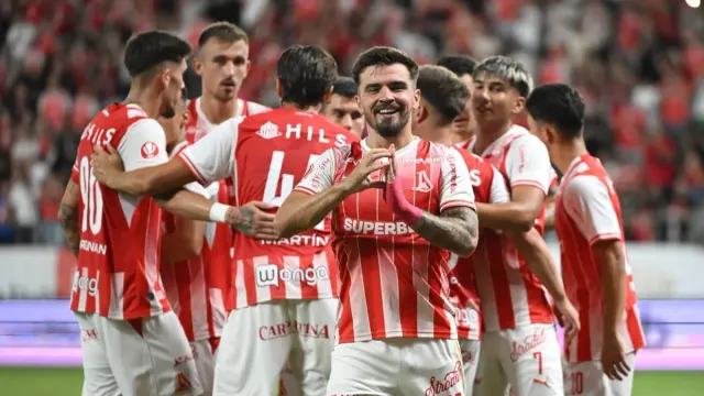 Fotbaliști în tricouri cu dungi roșii și albe, cu sponsorul Superbet, bucurându-se împreună pe teren