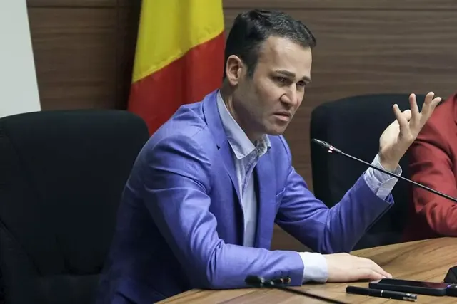 Bărbat în sacou albastru, gesticulând la o masă cu microfon, în fața tricolorului