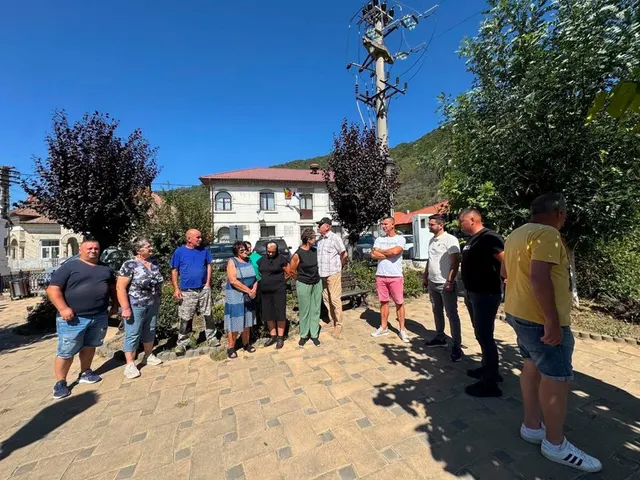 Grup de localnici adunați pe un trotuar, în fața unei clădiri, la poalele unor dealuri împădurite