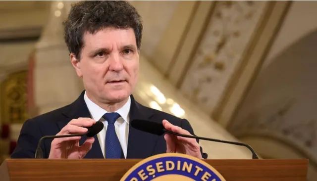 Bărbat cu părul creț, în costum și cravată albastră, la pupitrul cu stema Președintelui, într-un salon ornat