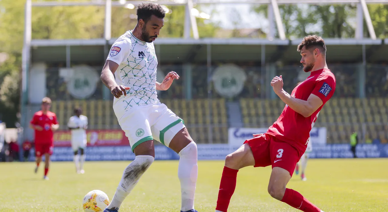 Fotbalist în echipament alb-verde, cu mingea la picior, urmărit de un adversar în roșu cu numărul 3