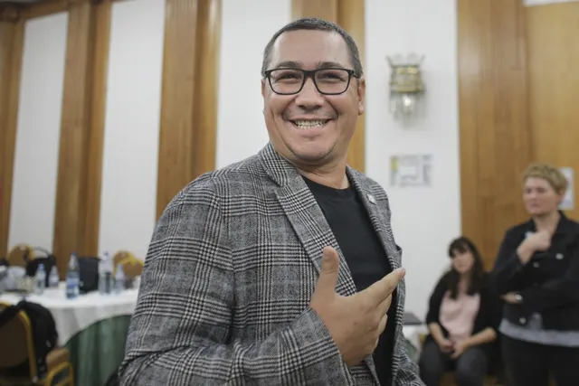 Bărbat zâmbitor cu ochelari, în sacou gri în carouri, gesticulând într-o sală cu lambriuri