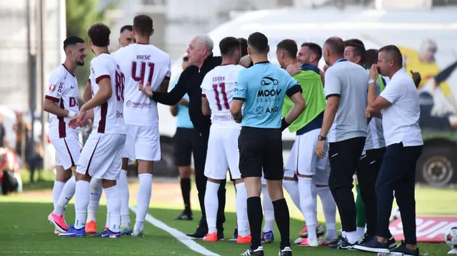 Fotbaliști în tricouri albe cu vișiniu și membri ai staffului, adunați în jurul arbitrului pe teren