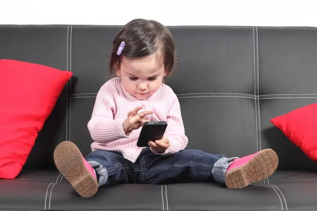 Fetiță mică în pulover roz, așezată pe o canapea, cu un telefon în mâini