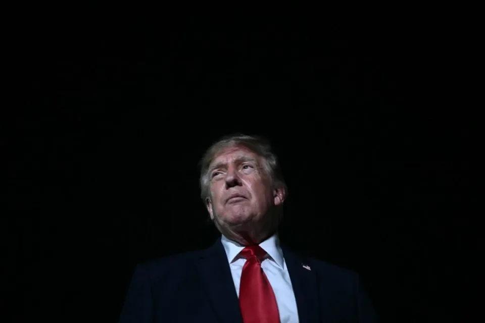 Donald Trump privind în sus, în costum închis și cravată roșie, pe un fundal negru