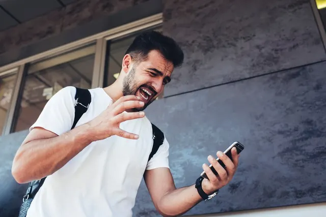 Tânăr cu barbă, în tricou alb și cu rucsac, zâmbind și gesticulând spre telefonul din mână