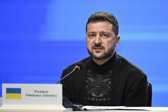 Volodimir Zelenski la o masă cu microfon, în spatele plăcuței cu numele său și drapelul Ucrainei