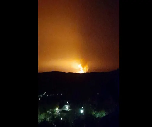 Flăcări și o lumină portocalie pe cerul nopții, deasupra unui deal, cu luminile unei localități dedesubt