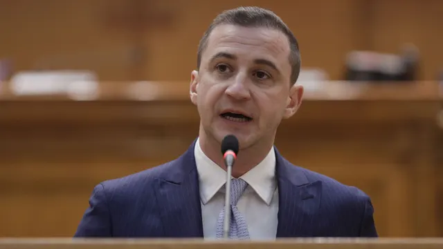 Bărbat tânăr în costum albastru vorbind la microfon de la tribuna din lemn a unei săli de plen