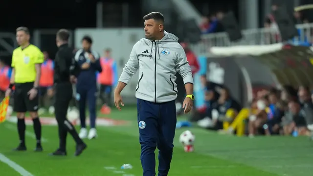 Bărbat în hanorac gri cu sigla clubului, mergând pe marginea terenului, cu arbitrul în spate