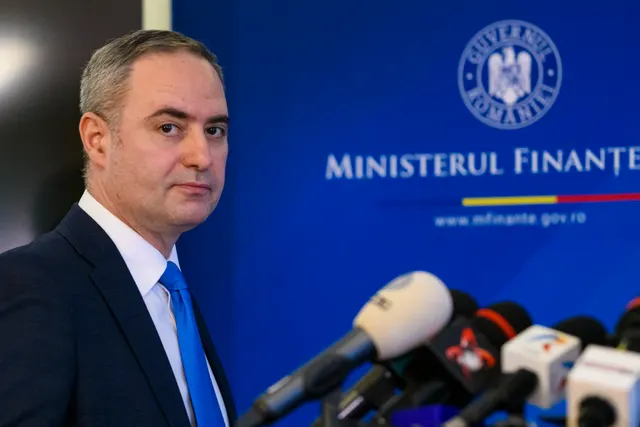 Bărbat în costum închis și cravată albastră, la microfoane, în fața panoului Ministerului Finanțelor