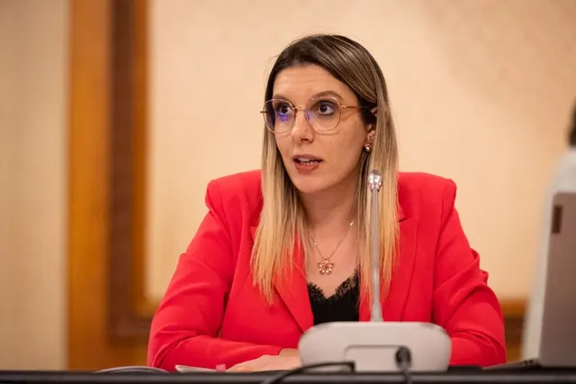 Femeie cu părul lung, blond, și ochelari, în sacou roșu, vorbind la o masă cu microfon