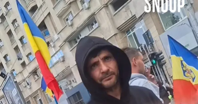 Bărbat cu glugă neagră în mijlocul unei mulțimi cu steaguri tricolore, pe o stradă cu blocuri