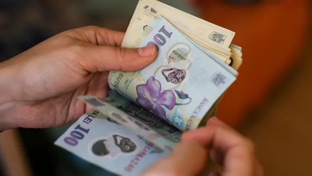 Mâini care numără un teanc de bancnote românești de 100 de lei, pe un fundal portocaliu estompat