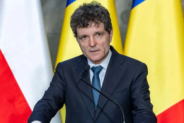 Bărbat cu părul creț vorbind la microfon, între drapelele Poloniei și României