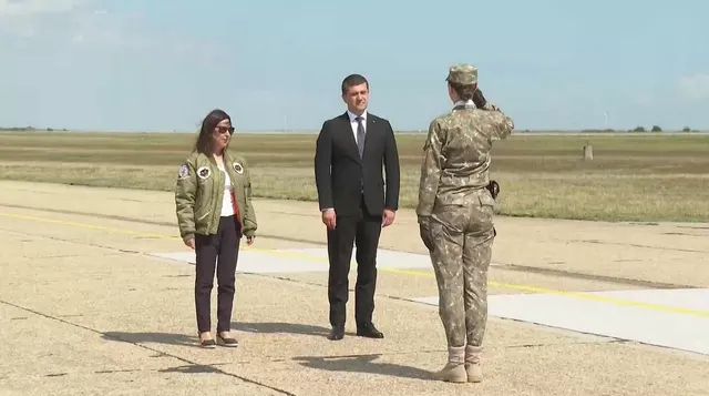 Captură de ecran: un bărbat în costum, o femeie în geacă kaki și o militară care salută, pe o pistă de aviație