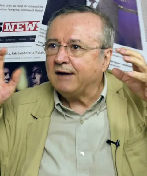 Ion Cristoiu, cu ochelari și sacou bej, vorbind în fața unui ecran cu titluri de știri
