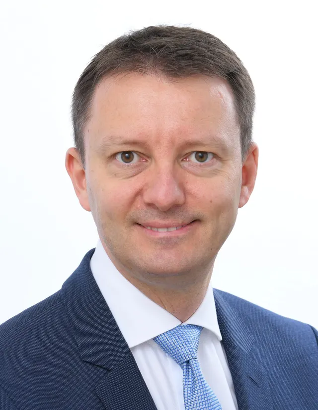 Portret al unui bărbat zâmbitor, în costum închis și cravată bleu, pe fundal alb