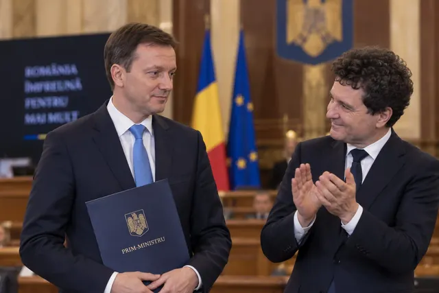 Doi bărbați în costume; unul ține o mapă albastră, celălalt aplaudă, lângă drapelele României și UE
