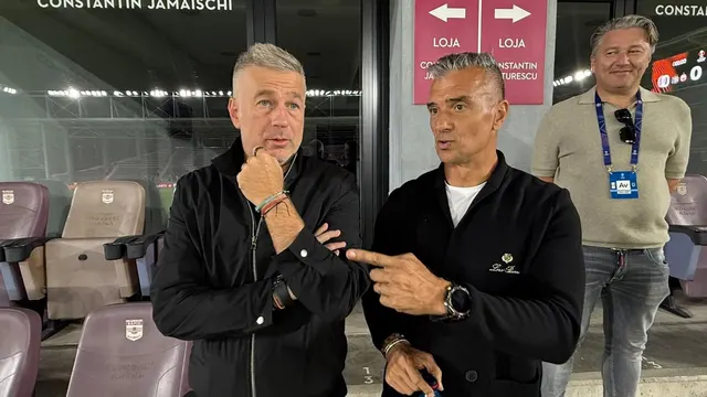 Trei bărbați discută într-o lojă de stadion, lângă scaune și o ușă cu inscripția „LOJA”