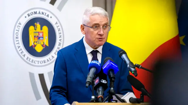 Bărbat cu părul alb și ochelari vorbind la microfoane, lângă sigla Autorității Electorale Permanente și tricolor