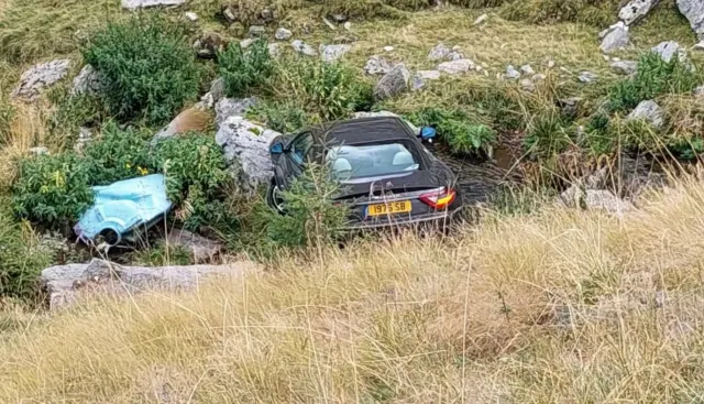 Mașină sport închisă la culoare, căzută într-o râpă printre stânci și iarbă uscată