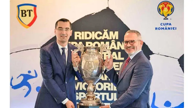 Doi bărbați în costume țin trofeul Cupei României, în fața unui panou cu siglele BT și Cupa României