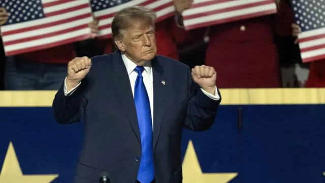 Bărbat în costum închis și cravată albastră, cu pumnii ridicați, în fața unor drapele americane