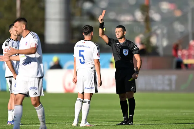 Arbitru cu brațul drept ridicat, lângă fotbaliști în echipament alb, unul cu numărul 5