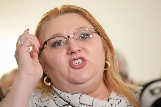 Femeie blondă cu ochelari, cu mâna la tâmplă, vorbind, în bluză cu imprimeu alb-negru