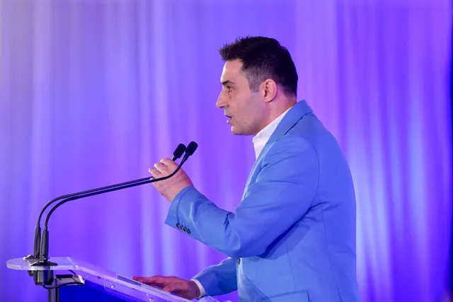 Bărbat în sacou albastru deschis vorbind la un pupitru cu microfon, pe un fundal luminat în violet