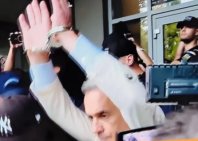 Bărbat cu părul cărunt ridicând mâinile împreunate, înconjurat de oameni cu camere de filmat