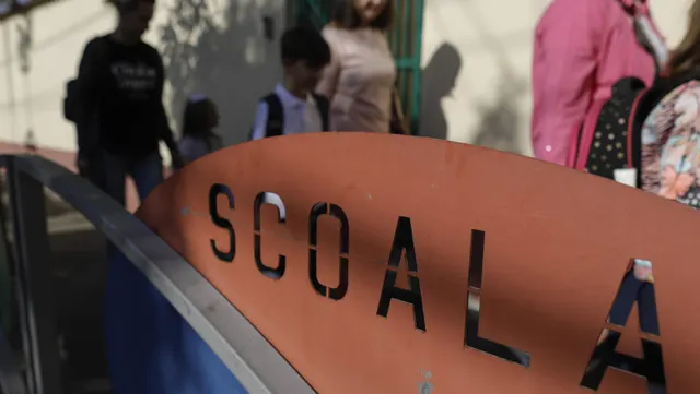 Inscripția „SCOALA” pe un panou curbat, cu tineri în fundal