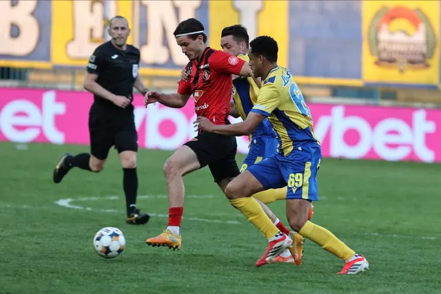 Fotbaliști în echipament roșu-negru și galben-albastru, în duel pentru minge, cu arbitrul în spate