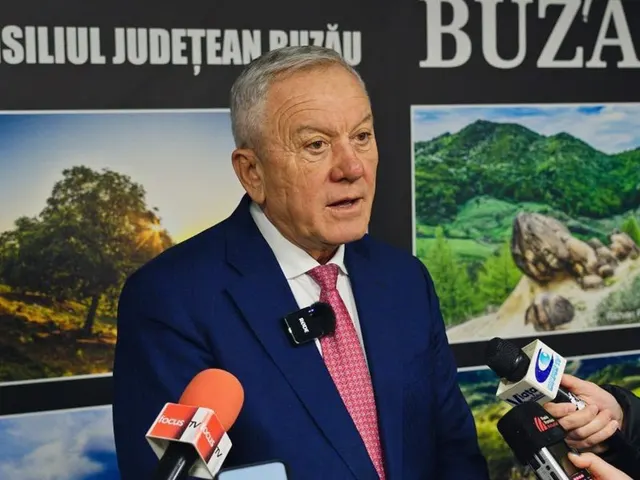 Bărbat cu părul alb, în costum albastru, vorbind la microfoane în fața unui panou al Consiliului Județean Buzău