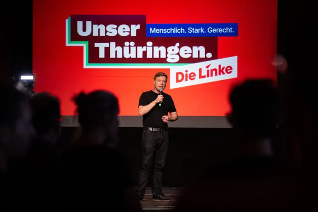 Bărbat cu microfon pe o scenă, în fața unui ecran roșu cu mesajul „Unser Thüringen” al partidului Die Linke