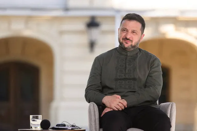 Bărbat în cămașă verde-închis cu broderie, așezat într-un fotoliu, zâmbind, în fața unei clădiri