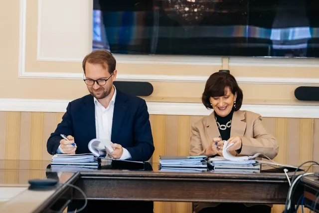 Un bărbat și o femeie semnând documente la o masă de lemn, într-o sală oficială