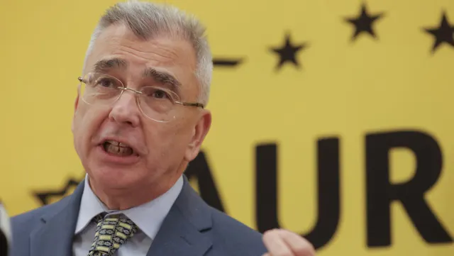 Bărbat cu ochelari și păr grizonat vorbind, în fața bannerului galben al AUR