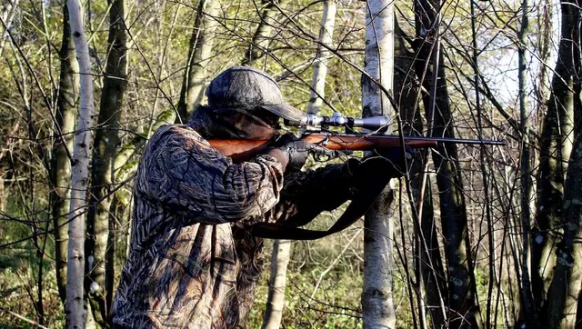 Vânător în haine de camuflaj ochind cu o pușcă cu lunetă, într-o pădure