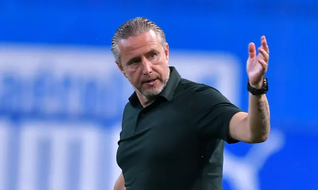 Bărbat cu părul grizonat, în tricou polo negru, gesticulând cu mâna ridicată, pe marginea terenului