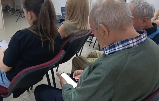 Bărbat în vârstă, în pulover verde, privind telefonul, printre participanți așezați într-o sală