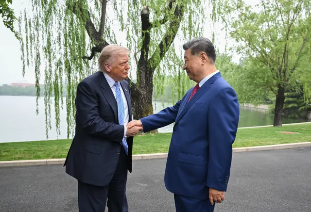 Donald Trump și Xi Jinping dând mâna în aer liber, lângă un lac cu sălcii