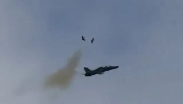 Avion militar cu reacție în zbor, cu o dâră de fum în spate și două obiecte mici deasupra lui