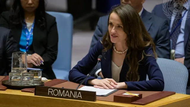 Femeie cu părul lung, șaten, la masa unei reuniuni internaționale, în spatele plăcuței „Romania”