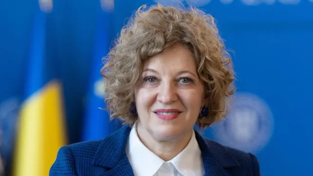 Femeie cu părul creț, șaten-blond, zâmbind, în sacou bleumarin, în fața unor drapele